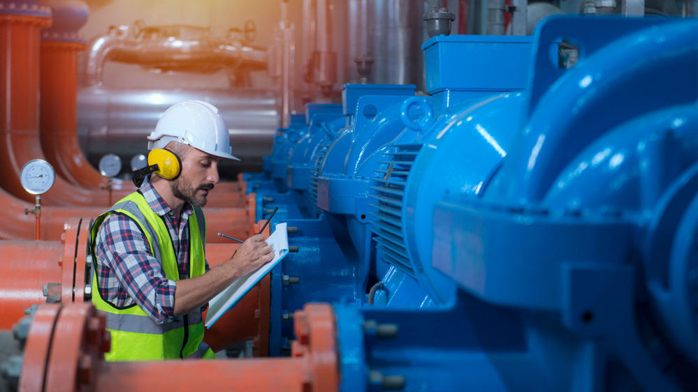 A mechanic writes something on a clipboard as he inspects an industrial centrifugal pump.